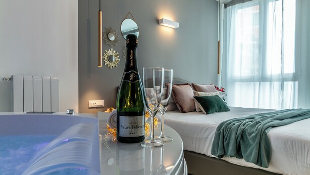 A modern bedroom with a bed, a bedside table with a bottle of Veuve Clicquot champagne and two glasses, a large window with sheer curtains, and a wall-mounted mirror. - 4 Star Suite SPA  (Bologna)