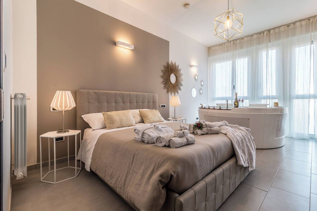A modern bedroom with a large bed, bedside lamps, a vanity, and a large window with curtains.