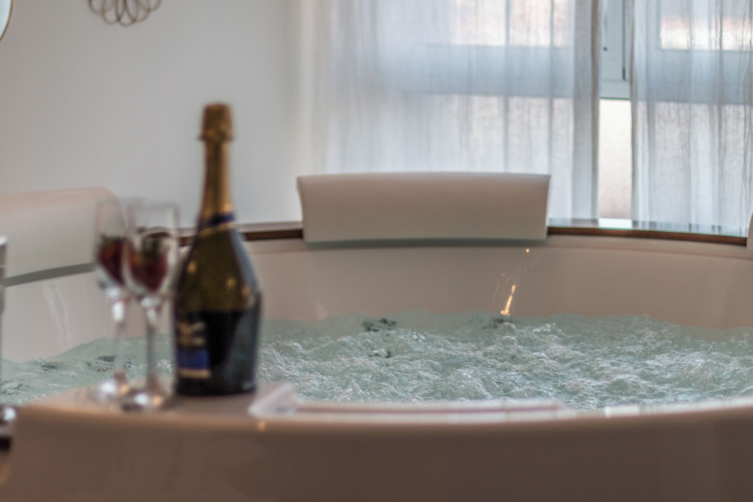 A table with a bottle of champagne and a glass, set against a window with sheer curtains.