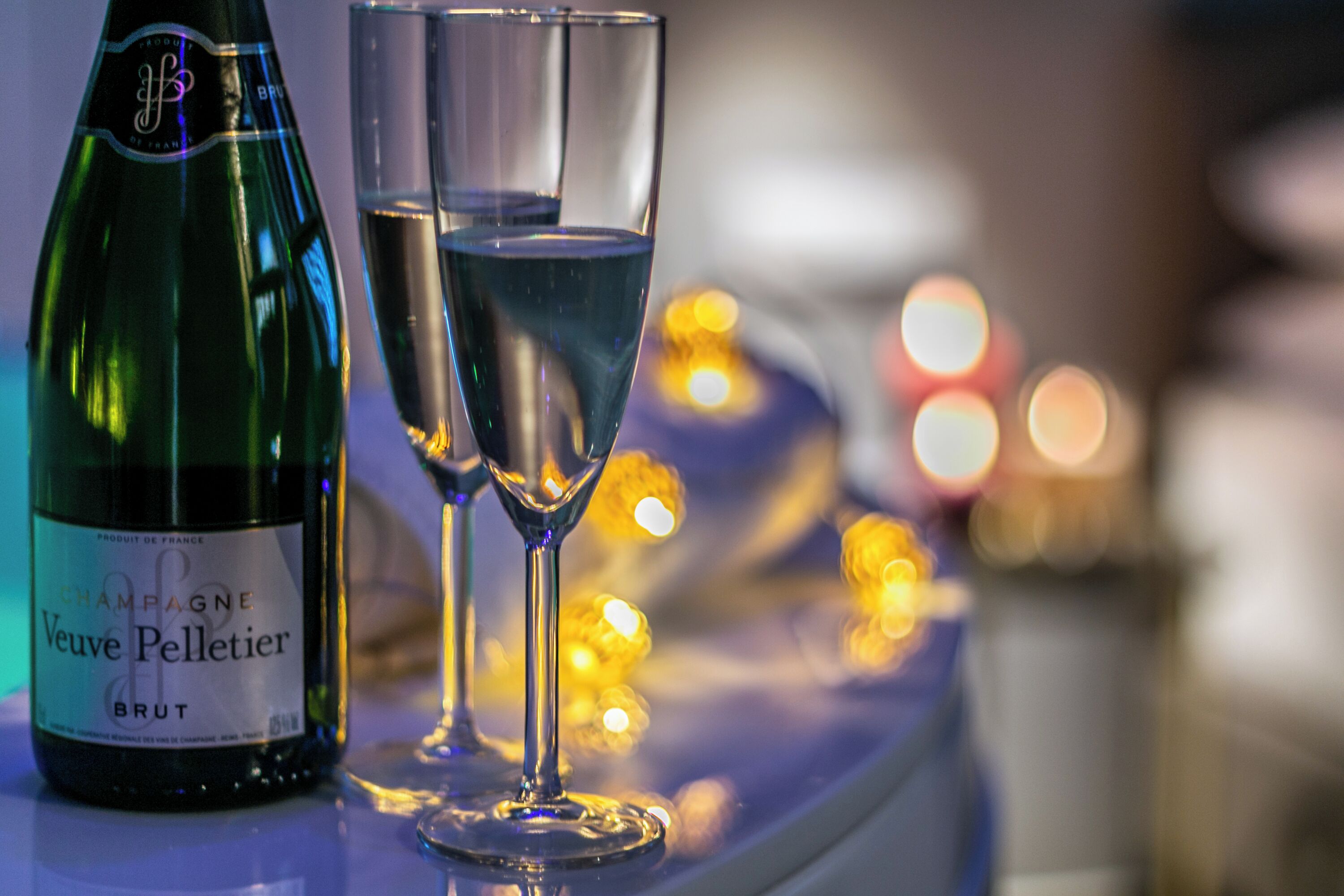 Eine Flasche Veuve Pellier Brut Champagner und zwei Gläser auf einer reflektierenden Oberfläche, im Hintergrund verschwommene Lichter.