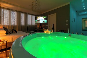 Una habitación con un jacuzzi grande iluminado en verde, una cama con almohadas, un televisor en la pared y una mesita con velas.