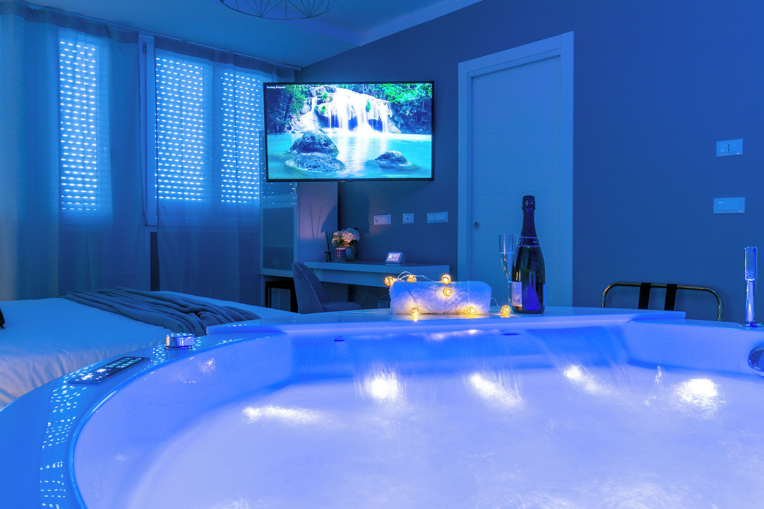 A modern hotel room with a large bathtub, a television mounted on the wall, and a small table with a bottle and glasses.
