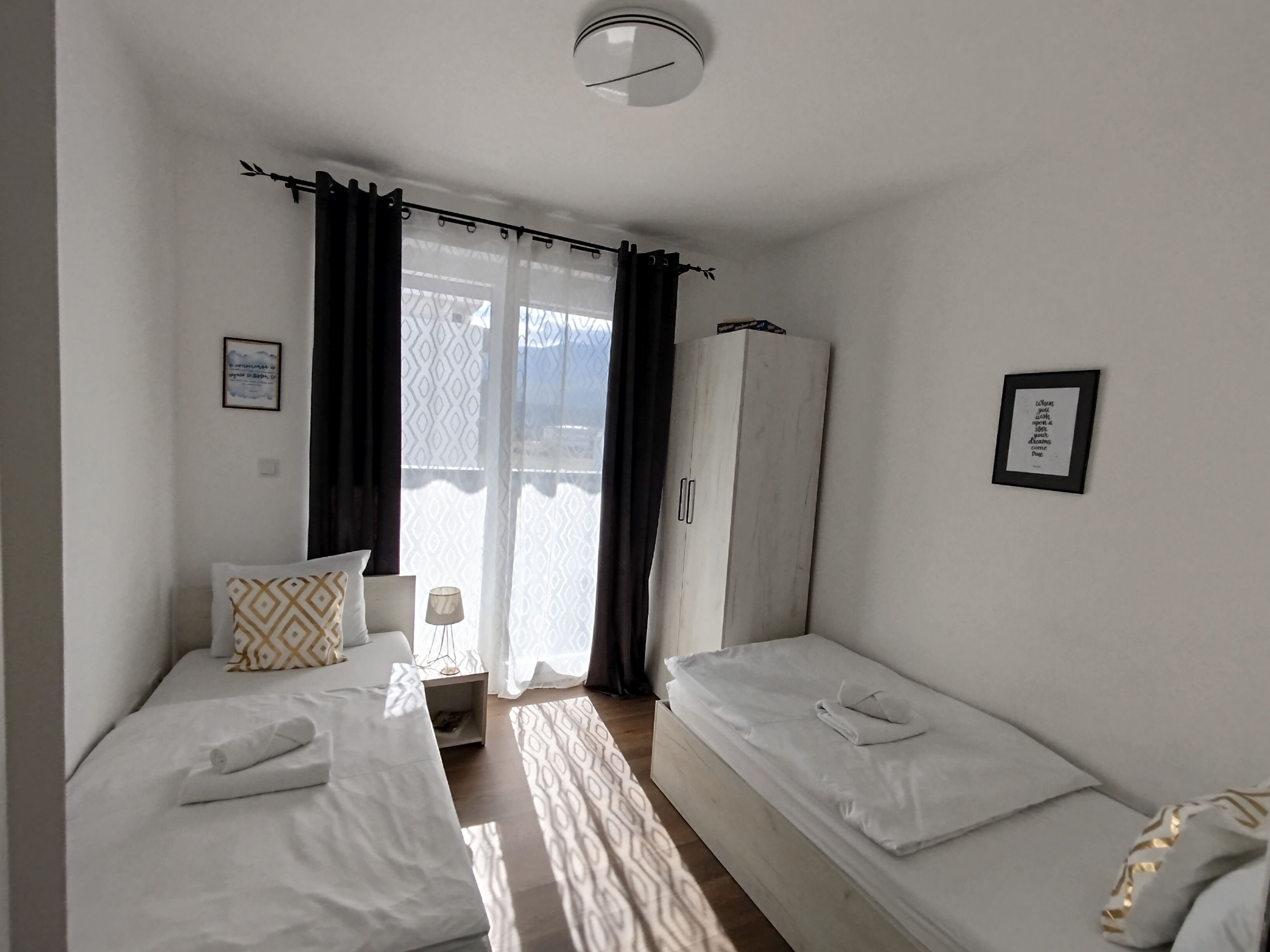 Habitación de hotel con dos camas individuales, un armario, una mesita de noche con lámpara y vistas a través de una ventana con cortinas.