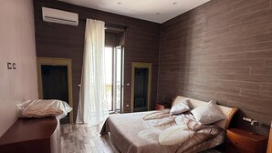 Un dormitorio con cama, mesita de noche y escritorio. Se ve una ventana con cortinas, una puerta y un equipo de aire acondicionado en la pared.