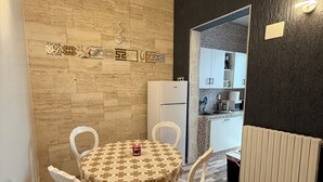 Un comedor con una mesa redonda cubierta con un mantel estampado, rodeada de sillas blancas. Una pequeña vela roja está colocada sobre la mesa. El ambiente tiene suelo de madera y una pared con un borde decorativo de azulejos.