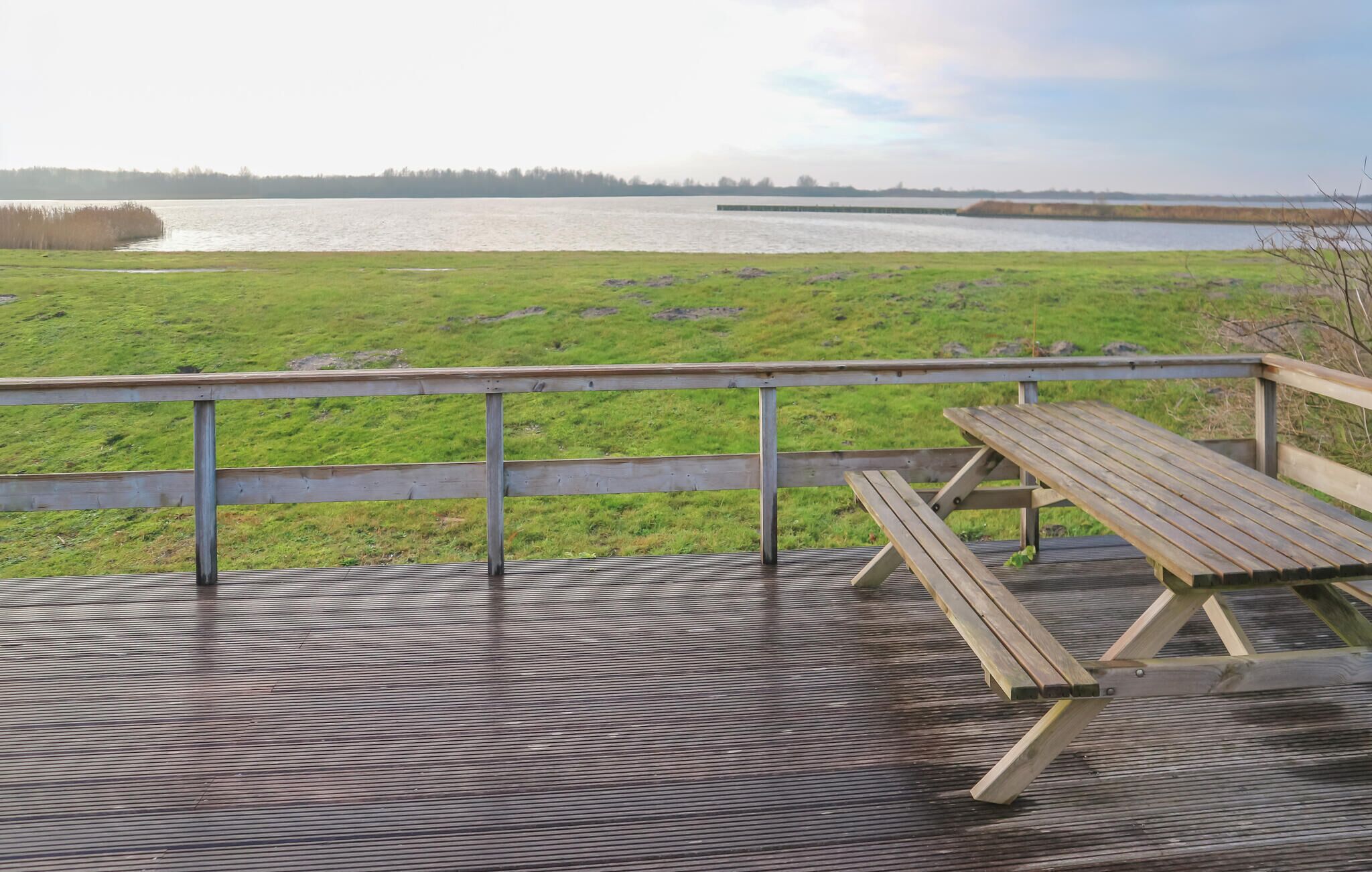 Een houten terras met een picknicktafel en een hekwerk, met uitzicht op een grasveld en een waterpartij.