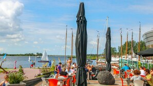 Ein Restaurant am Wasserufer mit Außenplätzen, Segelboote im Hintergrund und einem klaren Himmel.