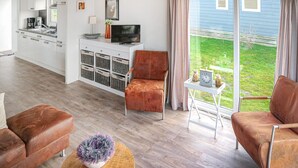 Ein modernes Wohnzimmer mit einer Couch, einem Sessel, einem kleinen Tisch mit einer Pflanze und einer Küchenzeile inklusive Fernseher und Schränken.