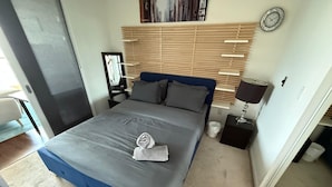 Une chambre à coucher avec un lit, une table de chevet avec une lampe, une horloge au mur et une fenêtre avec des stores.