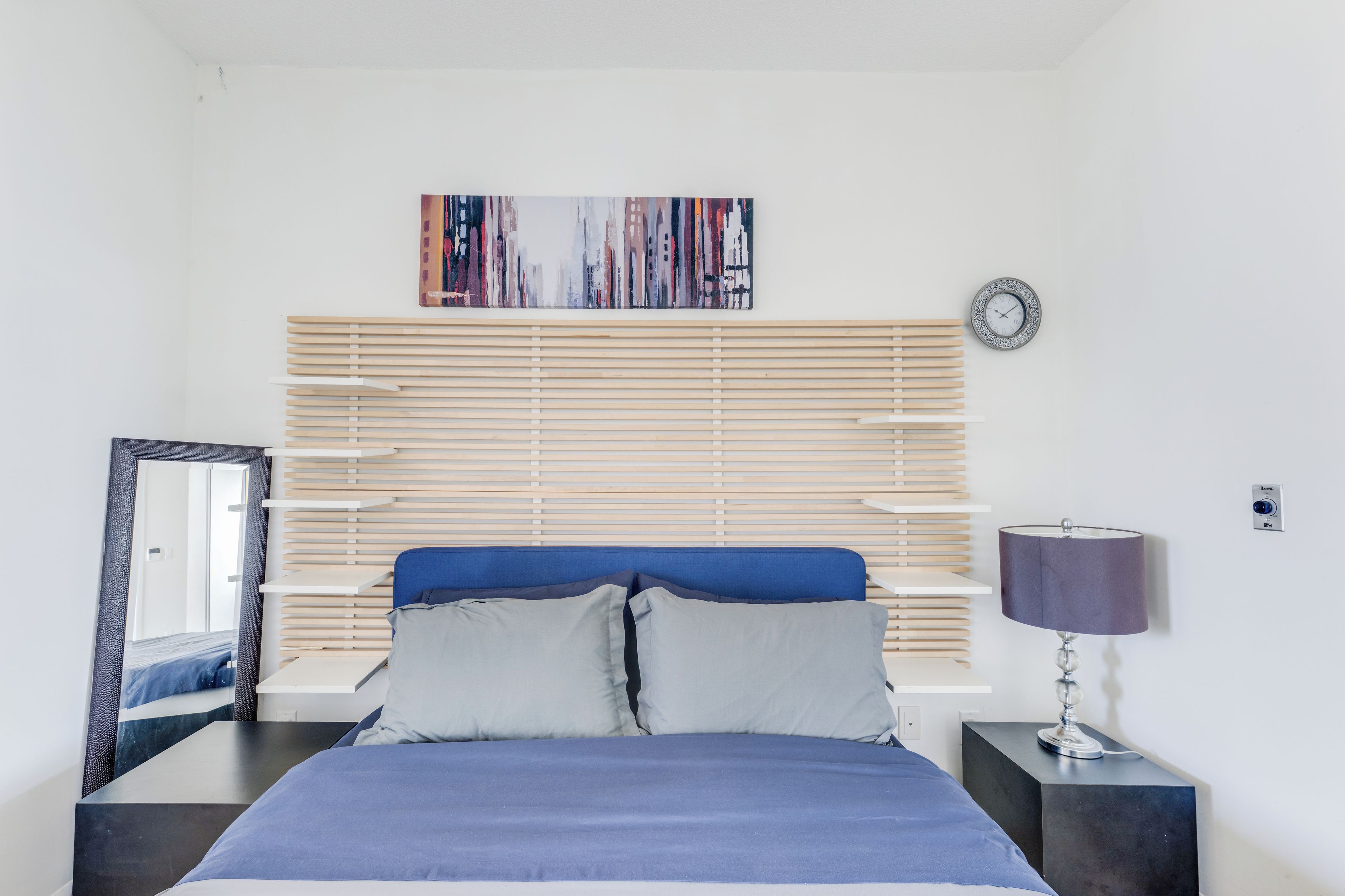 Een slaapkamer met een bed, nachtkastje, lamp, klok en schilderij aan de muur.