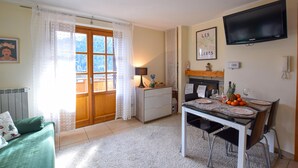 Uma sala de estar acolhedora com um sofá verde, uma mesa de jantar com cadeiras, uma TV na parede e uma janela com cortinas.