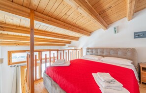 A bedroom with a red bedspread, wooden ceiling beams, a view through a window, and a nightstand with a lamp. - Lovely home in Carloforte with WiFi (Carloforte)