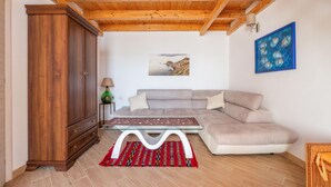 Ein Wohnzimmer mit Holzboden, einem beigefarbenen Sofa, einem Glastisch, einem roten Teppich mit Muster und einem Holzkasten. An der Wand hängt ein Gemälde.