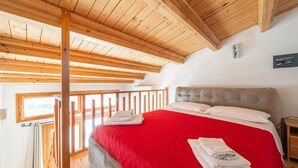 Ein Schlafzimmer mit Holzbalkendecke, einem großen Bett mit roter Bettwäsche und Blick ins Freie durch ein Fenster.