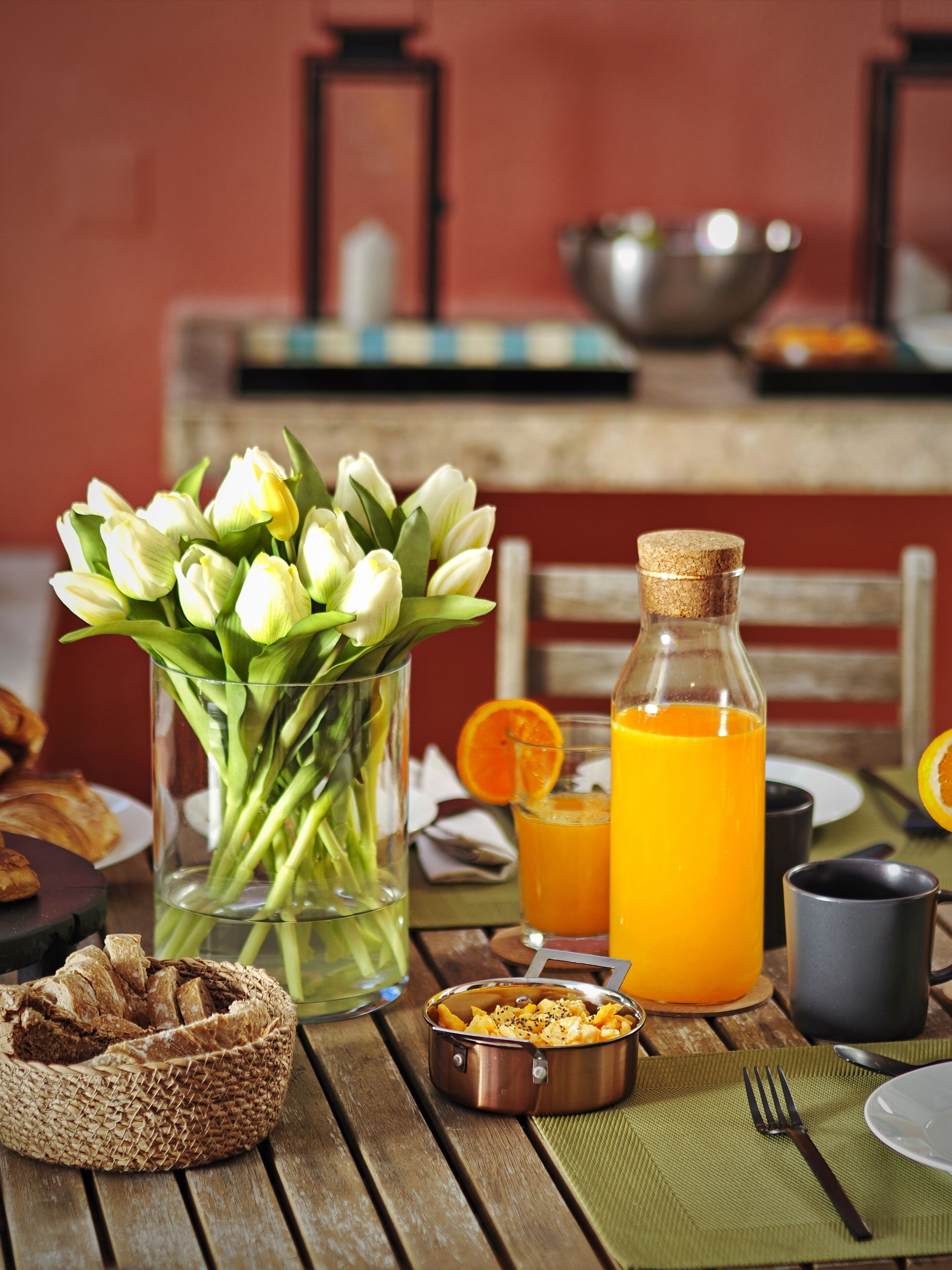 A breakfast setting with a vase of tulips, a bottle of orange juice, a bread basket, and a small bowl of cereal.