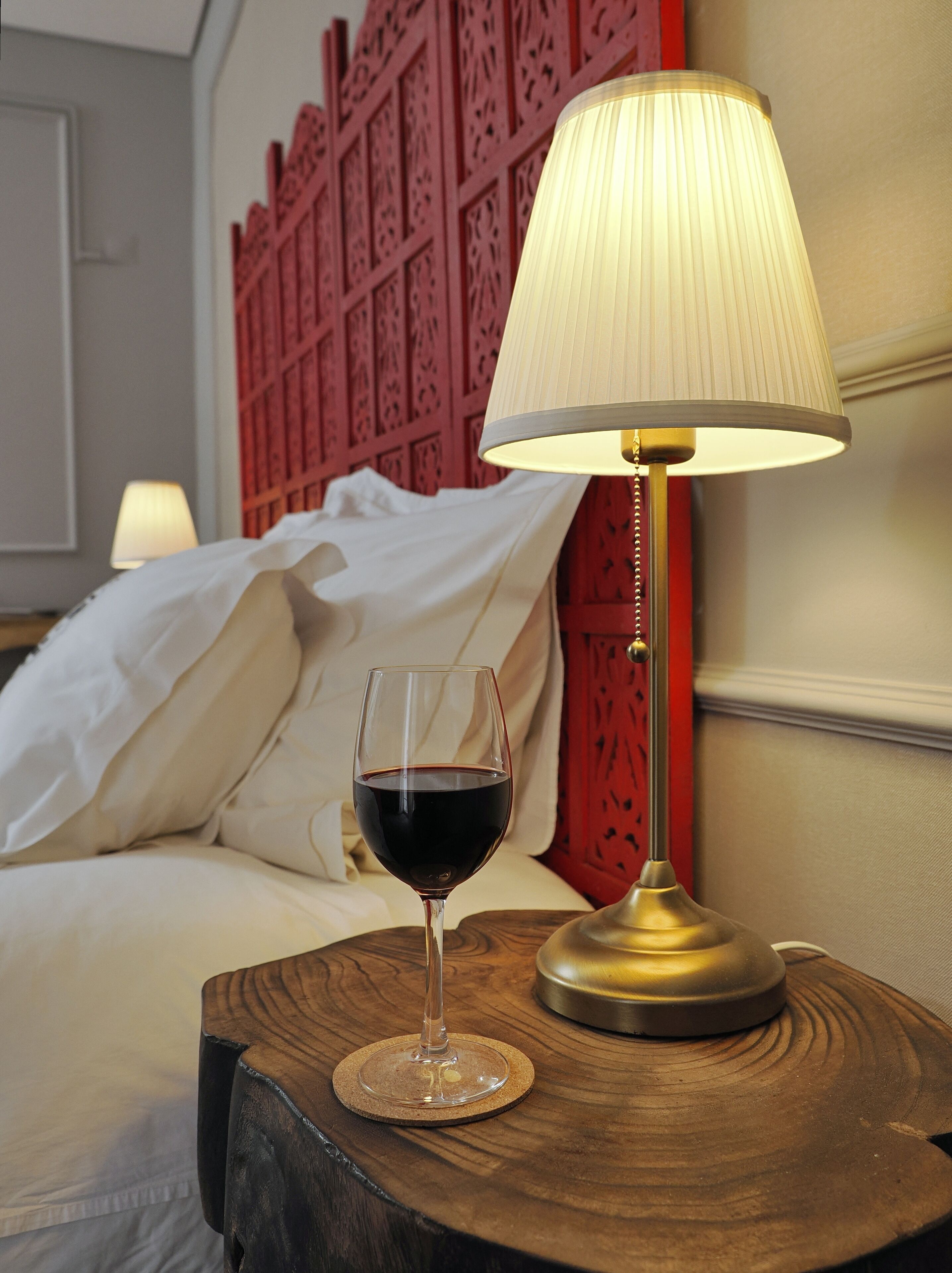 Un lit avec du linge de lit blanc, une tête de lit rouge et une table de chevet avec une lampe et un verre de vin rouge.