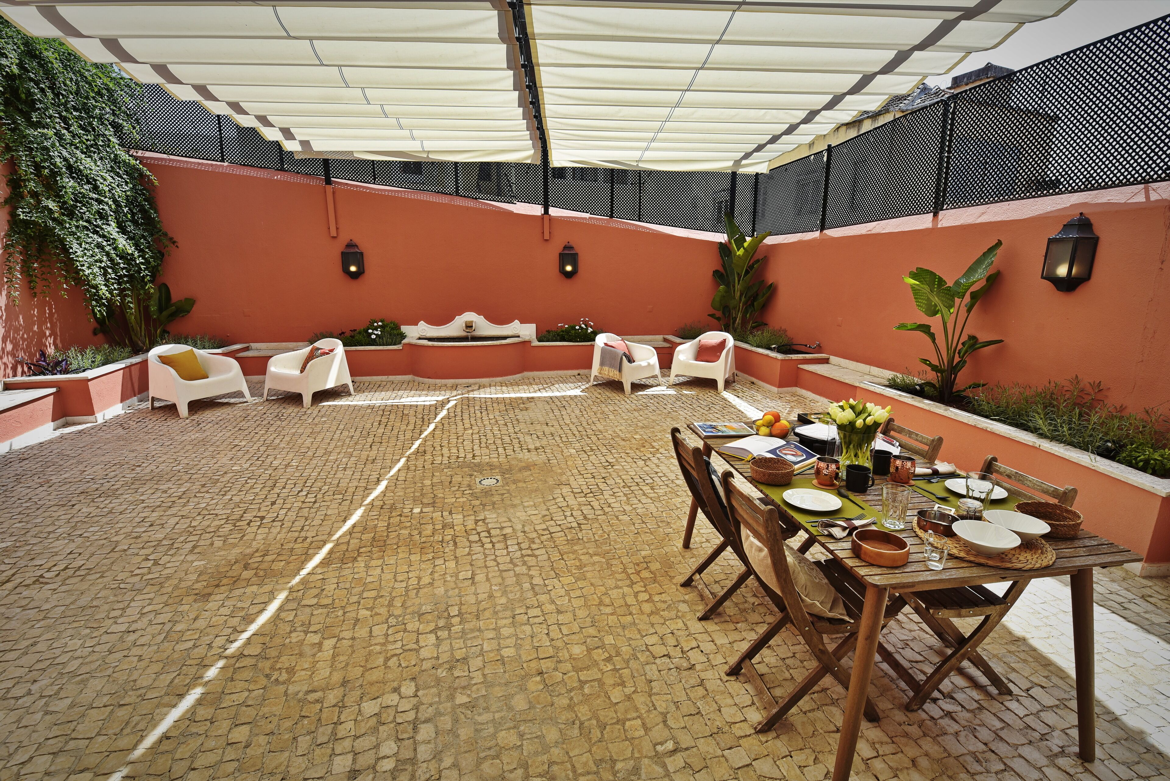 Un patio al aire libre con suelo de ladrillo, una zona de estar cubierta con sillas blancas y una mesa de comedor con platos y vasos.