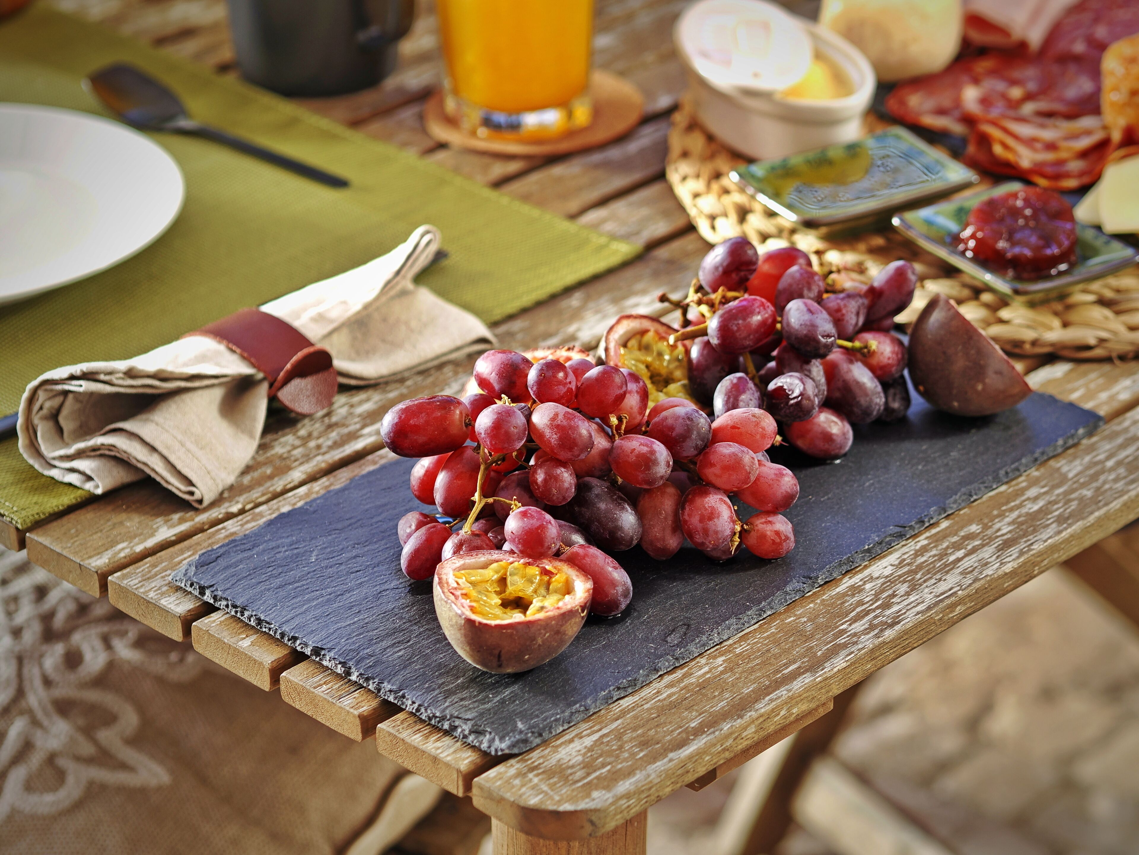 Una mesa de madera con una tabla de pizarra que sostiene un racimo de uvas rojas y media fruta, posiblemente una granada, con un mantel de servilleta verde y una cuchara al fondo.