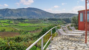 A terrace with stone flooring and white railings overlooking a valley with green fields and mountains.