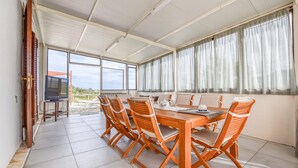 A spacious room with a large dining table and six chairs, large windows with curtains, and a television on a stand.