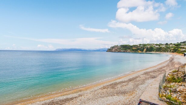 A coastal beach with clear water, pebbly shore, and houses on a hillside.