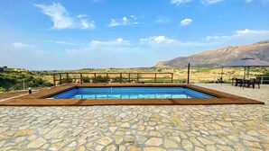 A swimming pool with a stone deck, wooden deck railing, and a view of distant hills and fields.