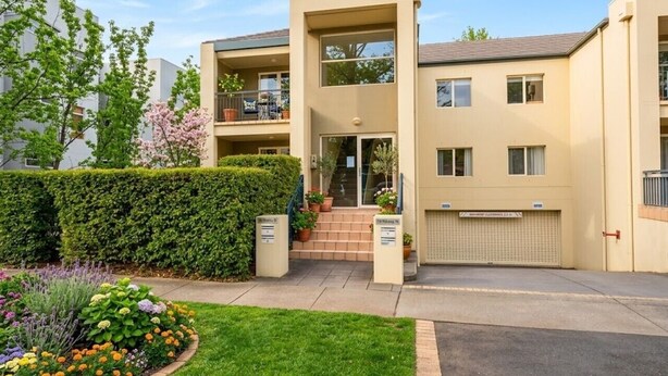 Een modern appartementencomplex met twee verdiepingen, een plat dak, een overdekte ingang en een kleine voortuin met verschillende planten en bloemen.
