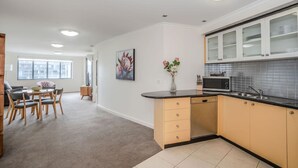 Una cocina moderna con encimera negra, armarios color beige y un microondas. Hay una zona de comedor con una mesa y sillas, y un cuadro en la pared.
