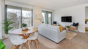 Ein modernes Wohnzimmer mit grauem Sofa, weißen Sesseln, einem runden Esstisch und einem an der Wand befestigten Fernseher.