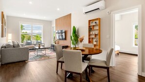 A modern living room with a dining area, large windows, and a flat-screen TV mounted on the wall. - New 2BR in DT Golden AC, Balcony, BBQ, Sleeps 6 (Golden)