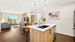 A modern kitchen with a central island, a dining area with orange chairs, and a living room with a grey sofa. - New 2BR in DT Golden AC, Balcony, BBQ, Sleeps 6 (Golden)