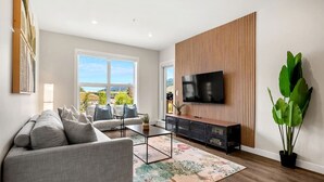 A modern living room with a sofa, coffee table, TV, and large windows offering a view of the outdoors. - New 2BR in DT Golden AC, Balcony, BBQ, Sleeps 6 (Golden)