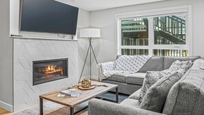 Ein modernes Wohnzimmer mit Kamin, einem flachbildfernseher darüber, einem grauen Sofa und einem großen Fenster mit Blick auf ein Gebäude.
