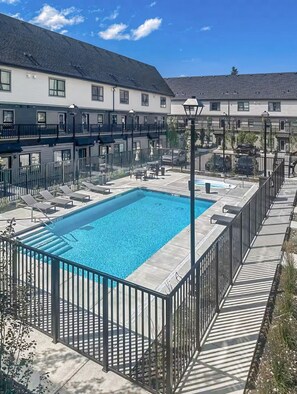 Una piscina rectangular envoltada per una tanca metàl·lica, amb un edifici de diversos pisos al fons.