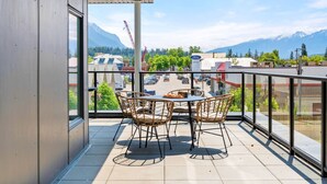 A balcony with a table and chairs, offering a view of a parking lot and mountains.
