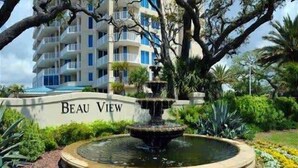 A fountain surrounded by a circular stone border with a sign reading "Beau View" in front of a multi-story building with balconies. - Beau View 1805 | The Perfect Long-Term Coastal Escape! (Biloxi)