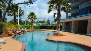 A swimming pool with a curved edge, surrounded by palm trees and a building with balconies. - Beau View 1805 | The Perfect Long-Term Coastal Escape! (Biloxi)