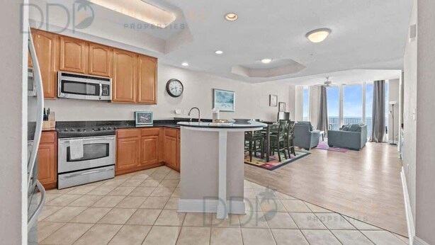 A modern kitchen with wooden cabinets, a central island, and a dining area with a view. - Beau View 1805 | The Perfect Long-Term Coastal Escape! (Biloxi)