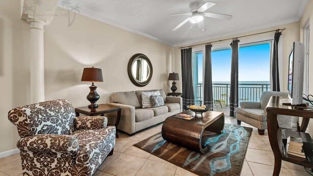 Uma sala de estar com sofá, poltrona, mesa de centro e televisão. Há uma varanda com vista para o oceano.