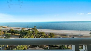 Vista costeira de uma varanda com vista para a praia, com uma estrada e carros estacionados ao primeiro plano.