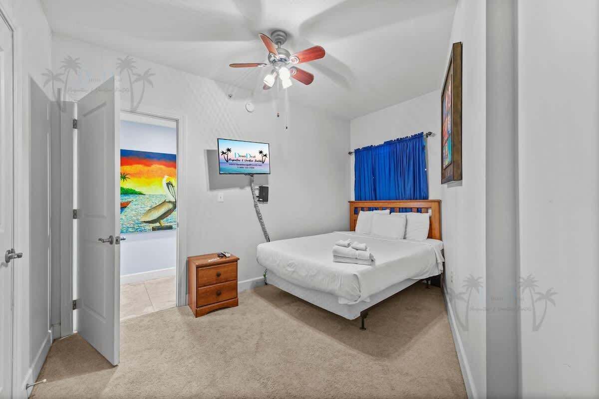 Un dormitorio con una cama, ventilador de techo, televisor, mesita de noche y una puerta.