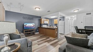 Ein modernes Wohnzimmer mit einem grauen Sofa, einem Couchtisch und einem Fernseher. Es gibt eine Küchenzeile mit Holzschränken und einer Mikrowelle.