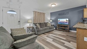 Ein Wohnzimmer mit grauem Sofa, gemusterten Kissen, einem Fernseher und einem Holzschrank.