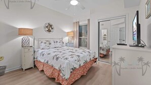 Ein Schlafzimmer mit Bett, Nachttischen, Deckenventilator, Spiegel und Fernseher.