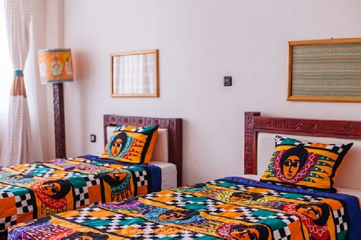 Una habitación con dos camas, cada una con colchas y almohadas de colores. Hay un cuadro enmarcado en la pared y una lámpara junto a las camas.