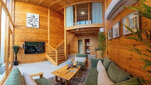 Interior de una cabaña de madera con una escalera de madera, un sofá, una mesa de centro y un televisor montado en la pared.