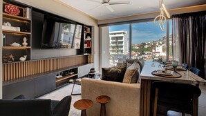 A modern living room with a large TV, a dining area with a table set for two, and a view of a cityscape through large windows. - Luxury Romantic Zone 1BR/2BA Condo + Rooftop Pool (Puerto Vallarta)