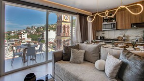 A modern living room with a large window, a cozy seating area, and a kitchen with wooden cabinets and a view of a clock tower. - Luxury Romantic Zone 1BR/2BA Condo + Rooftop Pool (Puerto Vallarta)