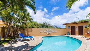 A swimming pool with a palm tree and lounge chairs, a sign reading "DU CH Villages", and a small building with doors. - Luxury Townhouse 6 Minutes from Beach & Casino (Oranjestad)