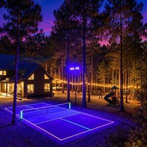 Una cabaña de madera con cancha de tenis, rodeada de árboles y luces colgantes.